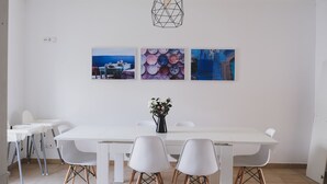 A modern dining area with a white table and chairs, a hanging light fixture, and three framed artworks on the wall.