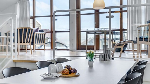 Ein moderner Essbereich mit einem weißen Tisch für zwei Personen, Meerblick durch große Fenster und ein Balkon mit Sitzgelegenheit im Freien.