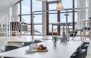 Un comedor moderno con una mesa blanca dispuesta para dos personas, vistas al mar a través de amplios ventanales y un balcón con mobiliario de exterior.
