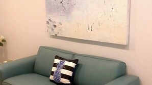 Un divano color petrolio con un cuscino a forma di ananas, un tavolino da caffè trasparente, una sveglia vintage e un quadro appeso al muro.