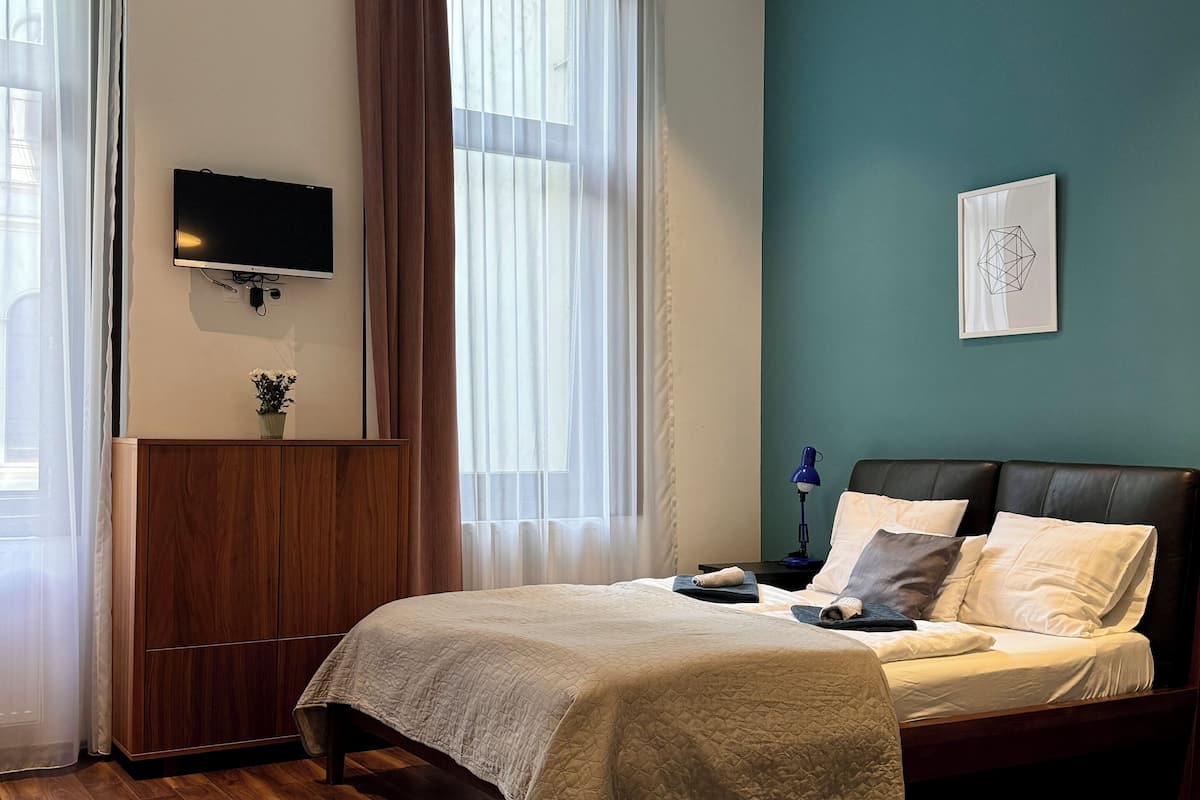 Un dormitorio con cama, televisión montada en la pared, ventana con cortinas y un cuadro en la pared.