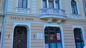 Um edifício com detalhes arquitetônicos elaborados, incluindo janelas em arco e varandas decorativas. O andar térreo tem uma placa com a inscrição "Katie's House" e vários avisos publicando serviços como armazenamento de bagagem e traslado para o aeroporto.