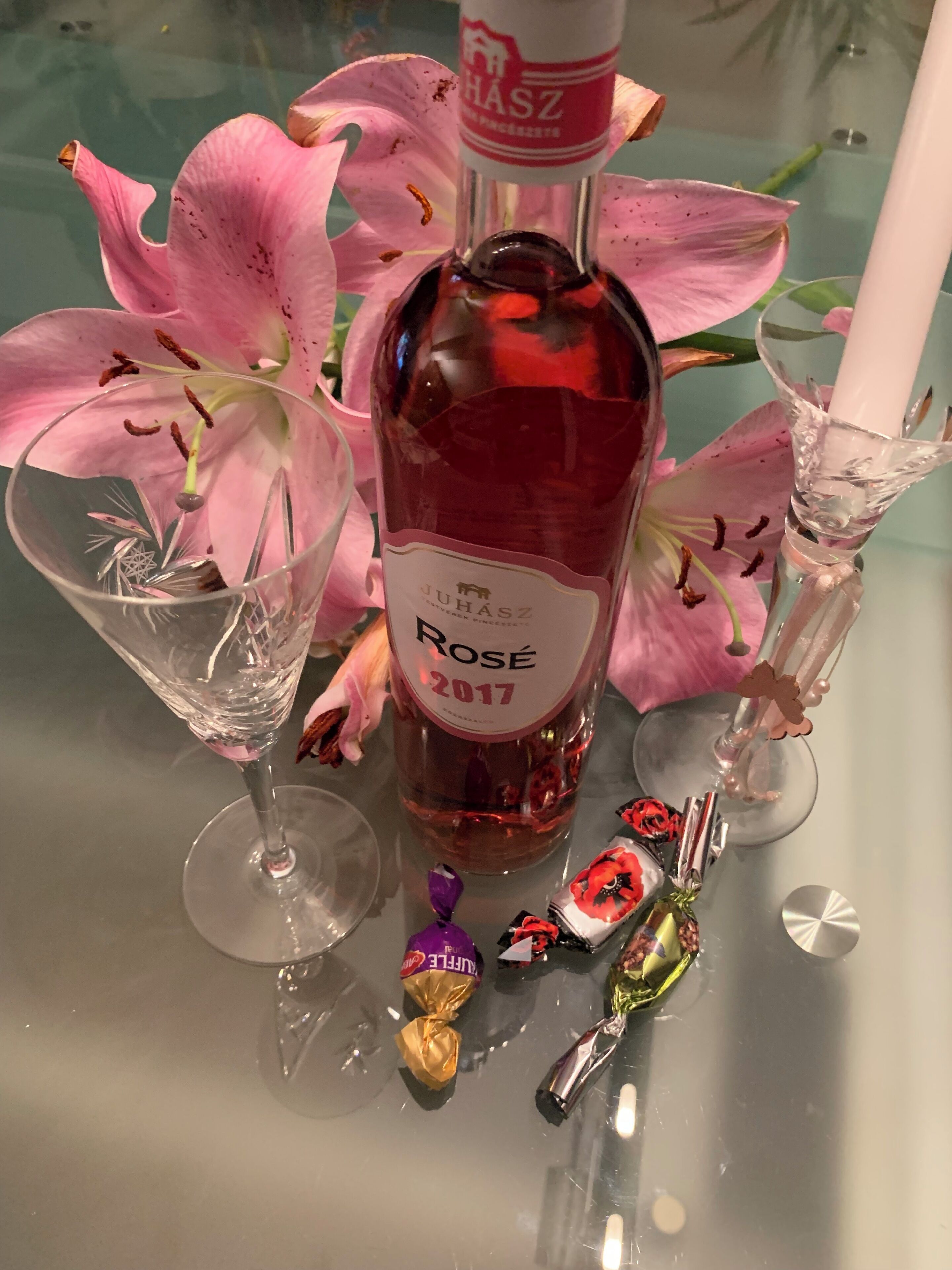 A bottle of wine, a glass, and a candle on a table with flowers and chocolates.