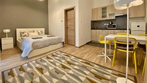 Quarto moderno com cama, mesa de cabeceira e criado-mudo. Ao lado do quarto, encontra-se uma cozinha com área de refeições, incluindo uma mesa e cadeiras.