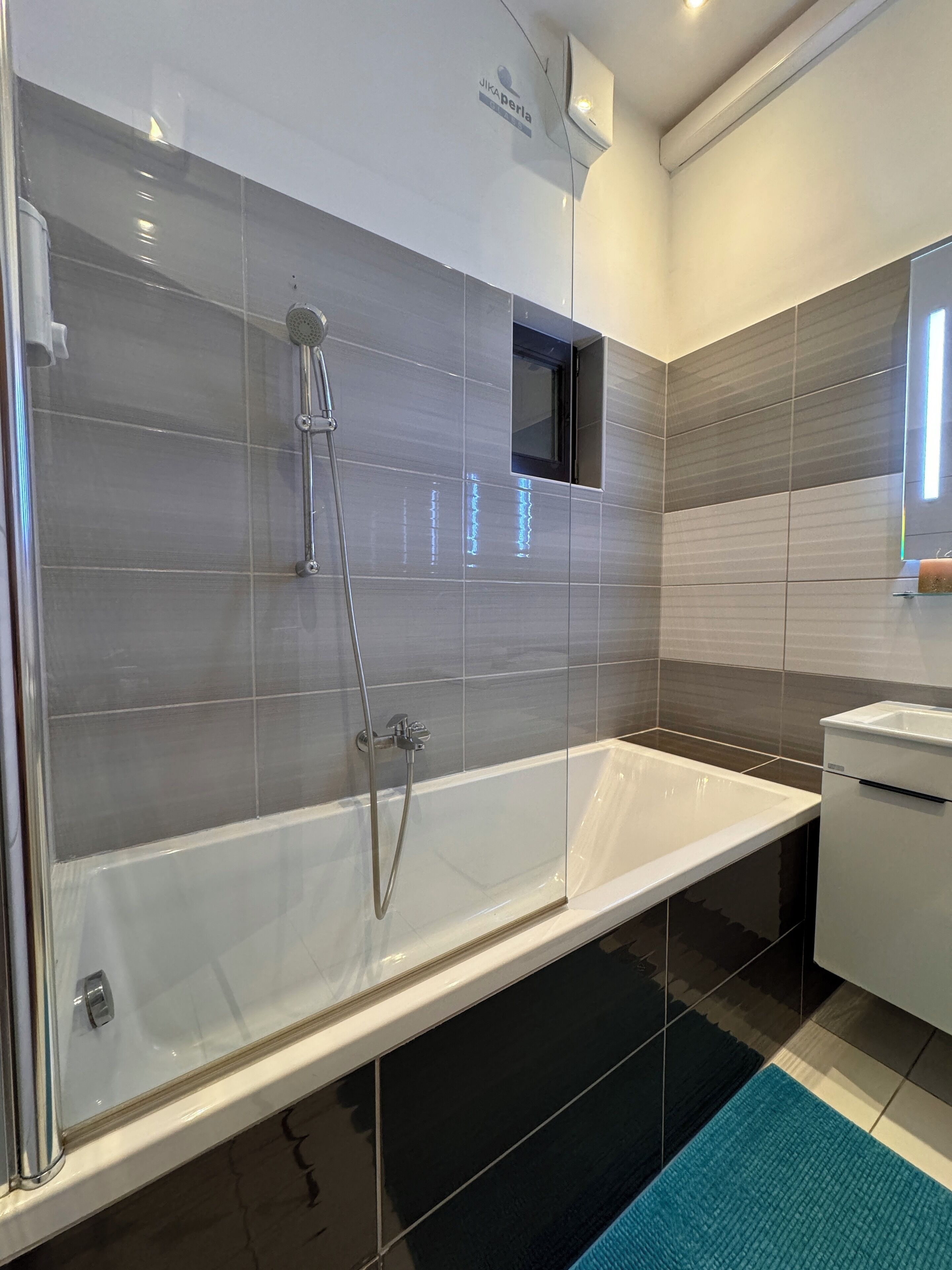 A modern bathroom with a glass-enclosed bathtub, a handheld showerhead, and a small window above the tub.
