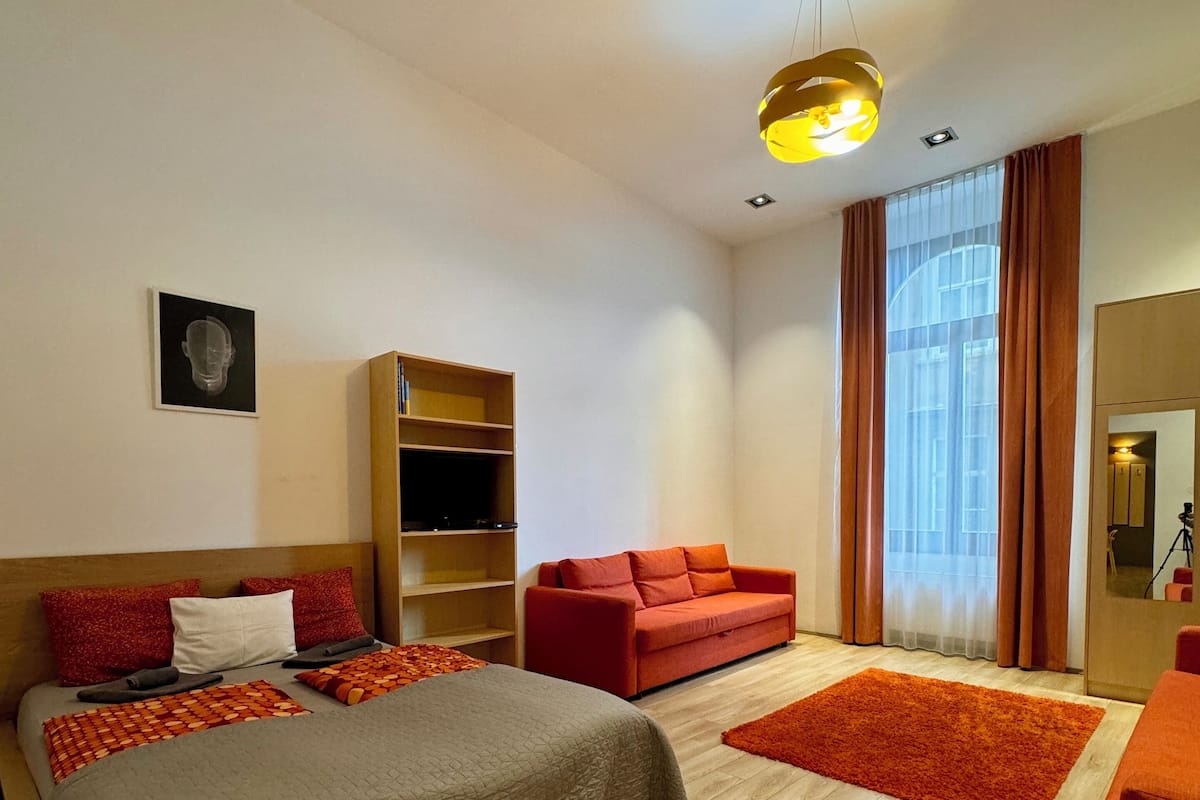 Una habitación de hotel moderna con una cama grande, un sofá rojo, suelo de madera y un amplio ventanal con cortinas.