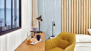 Une chambre d’hôtel avec un bureau en bois, deux lampes, un fauteuil jaune et un lit avec du linge de lit blanc.