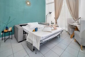 A compact hotel room with a bed, bedside table, lamp, and a dining area with chairs and a table set for two.