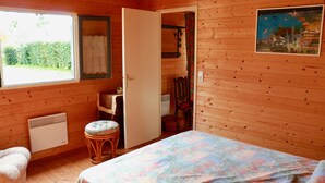 A wooden cabin room with a bed, a chair, a table, a window, and a door leading to another room. - Welcome to Rue des oiseaux (Névez)