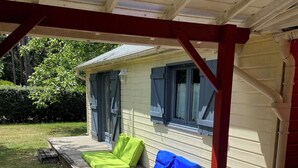 A covered porch with a wooden deck, a small house with blue shutters, and green and blue cushions on the deck.