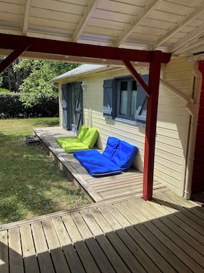 A covered porch with a wooden deck, a small house with blue shutters, and green and blue cushions on the deck.