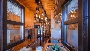 Un interior de madera con una zona de cocina, una mesa de comedor con una vela y un cuenco de plantas, y una ventana con cortinas de encaje.