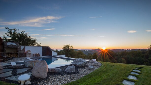 Un área de piscina moderna al aire libre con un sendero de piedra que conduce a un espacio de descanso, rodeado de un césped cuidado y vistas a la puesta de sol sobre un paisaje montañoso.