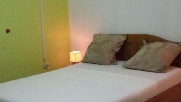 A single bed with a wooden frame and a white mattress, accompanied by two pillows. The room has a green wall on one side and a white wall on the other. - Motel Jupiter (Sangmélima)