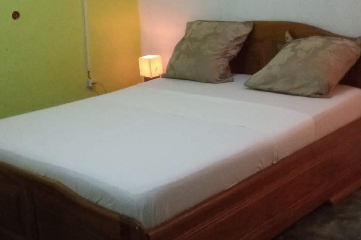 Una cama individual con estructura de madera y colchón blanco, acompañada por dos almohadas. La habitación tiene una pared verde en un lado y una pared blanca en el otro.