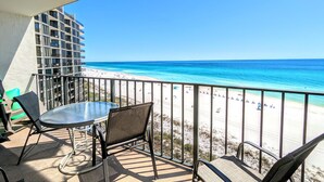 A balcony with a table and chairs, overlooking a beach with clear blue waters and a sandy shore. - Exquisite Deluxe Vacation 7th floor Tower II (Panama City Beach)