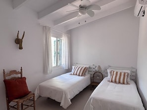 A room with two beds, a ceiling fan, a window with curtains, and a decorative wall piece.