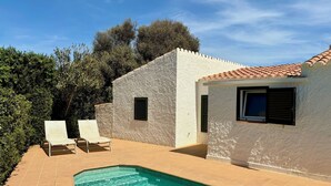 Zona junto a la piscina con una piscina azul cristalina, dos sillones de descanso y un edificio blanco con tejado de baldosas.