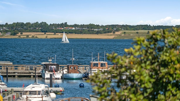 A marina with various boats docked, a sailboat in the distance, and a view of a residential area across the water. - Cozy home in Stubbekøbing with kitchen (Stubbekøbing)
