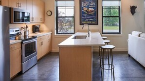 Une cuisine moderne avec des meubles en bois, des appareils électroménagers en acier inoxydable, un îlot central doté de tabourets et une grande fenêtre offrant une vue sur l’extérieur.