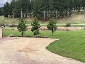 Surrounded by a dense forest and a hill, this property features a gravel driveway, grassy area, and a wooden fence.