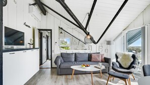 Un salon moderne avec un canapé gris, une table basse ronde et un fauteuil moelleux. Un téléviseur à écran plat se trouve à gauche et une grande fenêtre à droite.