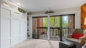 Een ruime kamer met een schuifdeur naar een terras, een zithoek met een bank en een voetenbankje, en een witte inbouwkast.