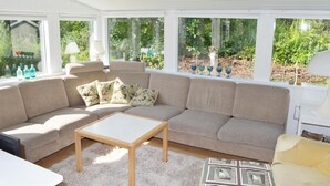 Una sala de estar acogedora con un sofá seccional color beige, una mesa de centro de madera y vistas a zonas verdes a través de amplios ventanales.