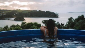 A person in a blue pool with a glass of red wine, overlooking a body of water and land. - Ocean View (Waiheke Island)