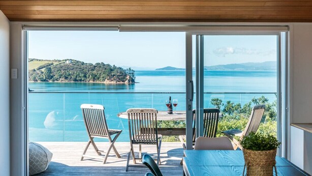 A balcony with a view of a lake and hills, wooden furniture, and a table with a plant. - Ocean View (Waiheke Island)