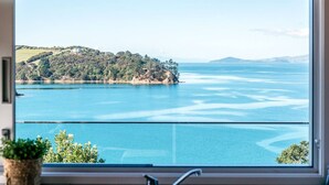 A view through a window of a coastal landscape with a body of water, a small island, and distant hills. - Ocean View (Waiheke Island)
