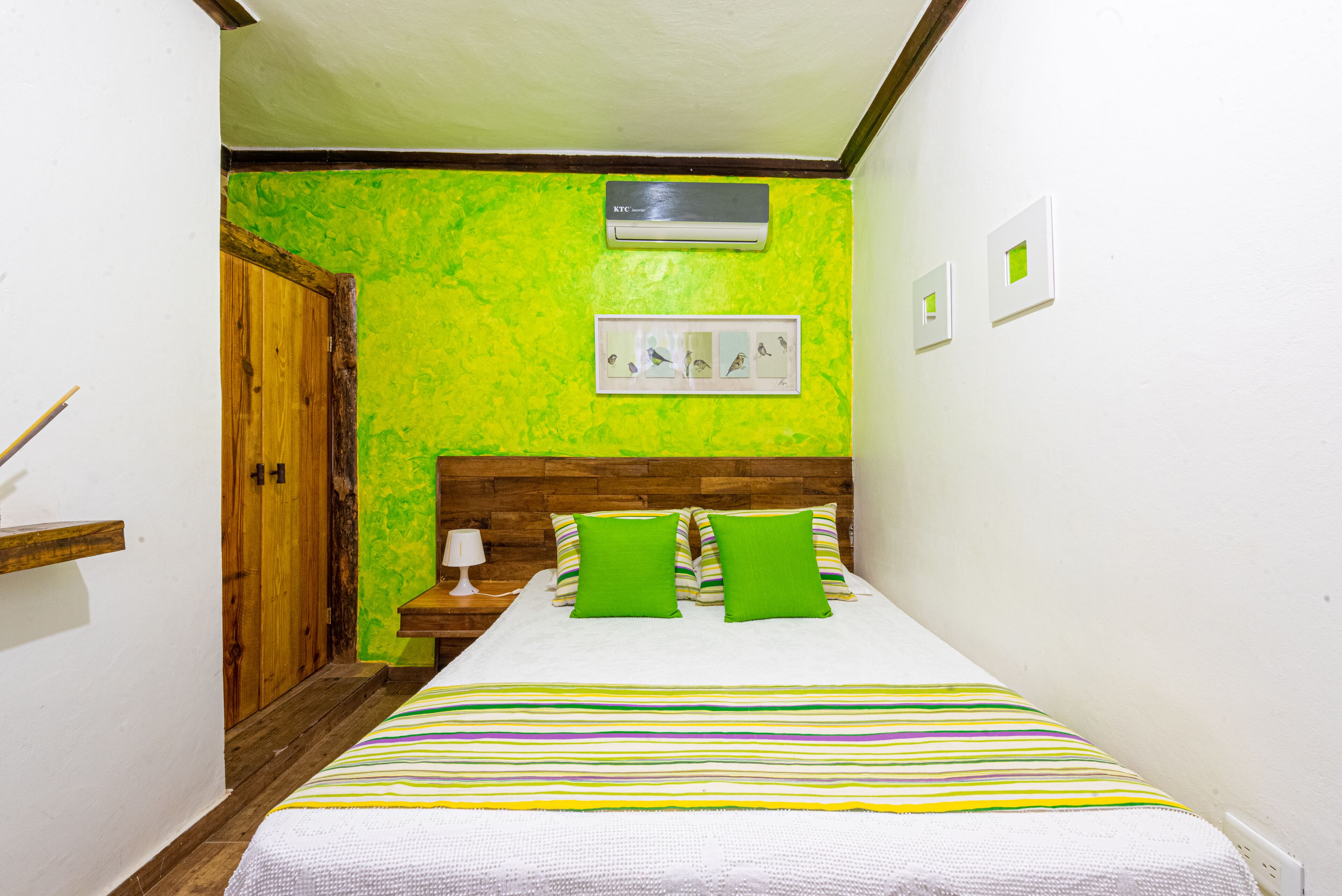 A bedroom with a wooden headboard, a bed with green pillows, a striped bedspread, a nightstand with a lamp, and a wall-mounted air conditioner.