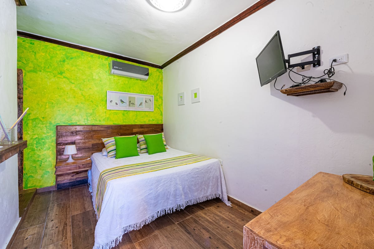 A room with a bed, a wooden nightstand, a wall-mounted TV, and a small table.