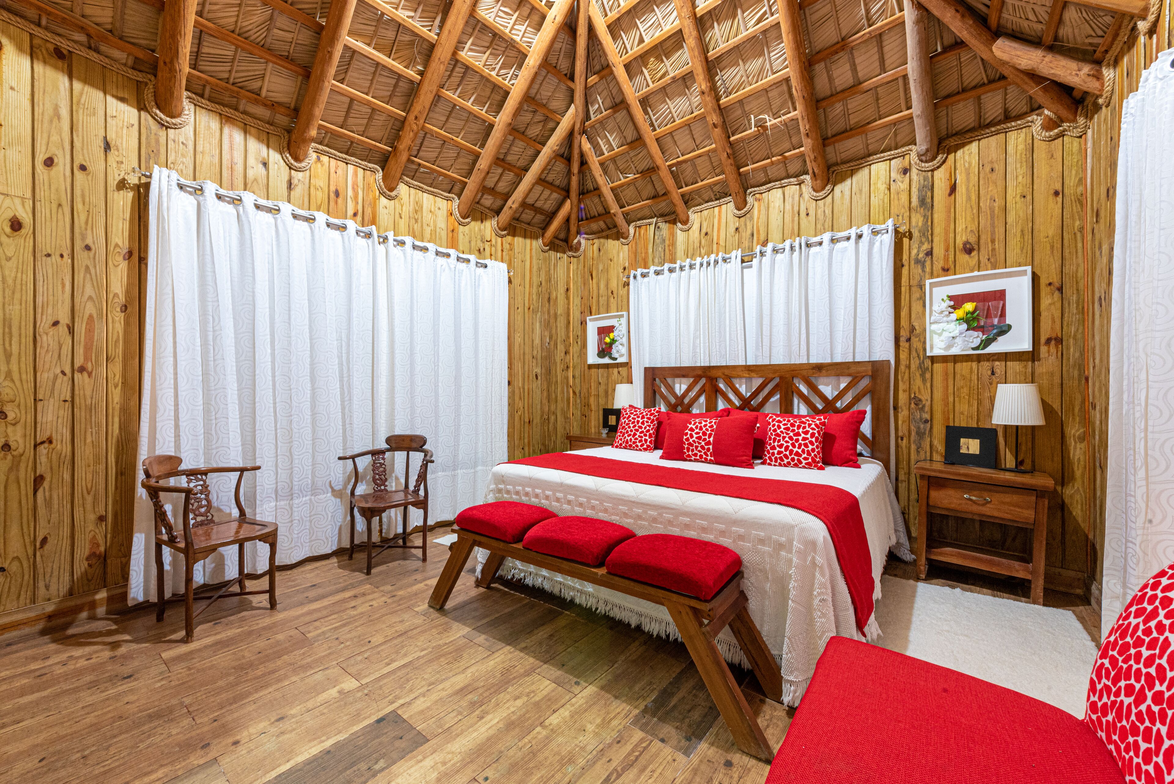 Una habitación con paneles de madera, una cama, dos sillas, un banco y una mesita de noche con una lámpara.