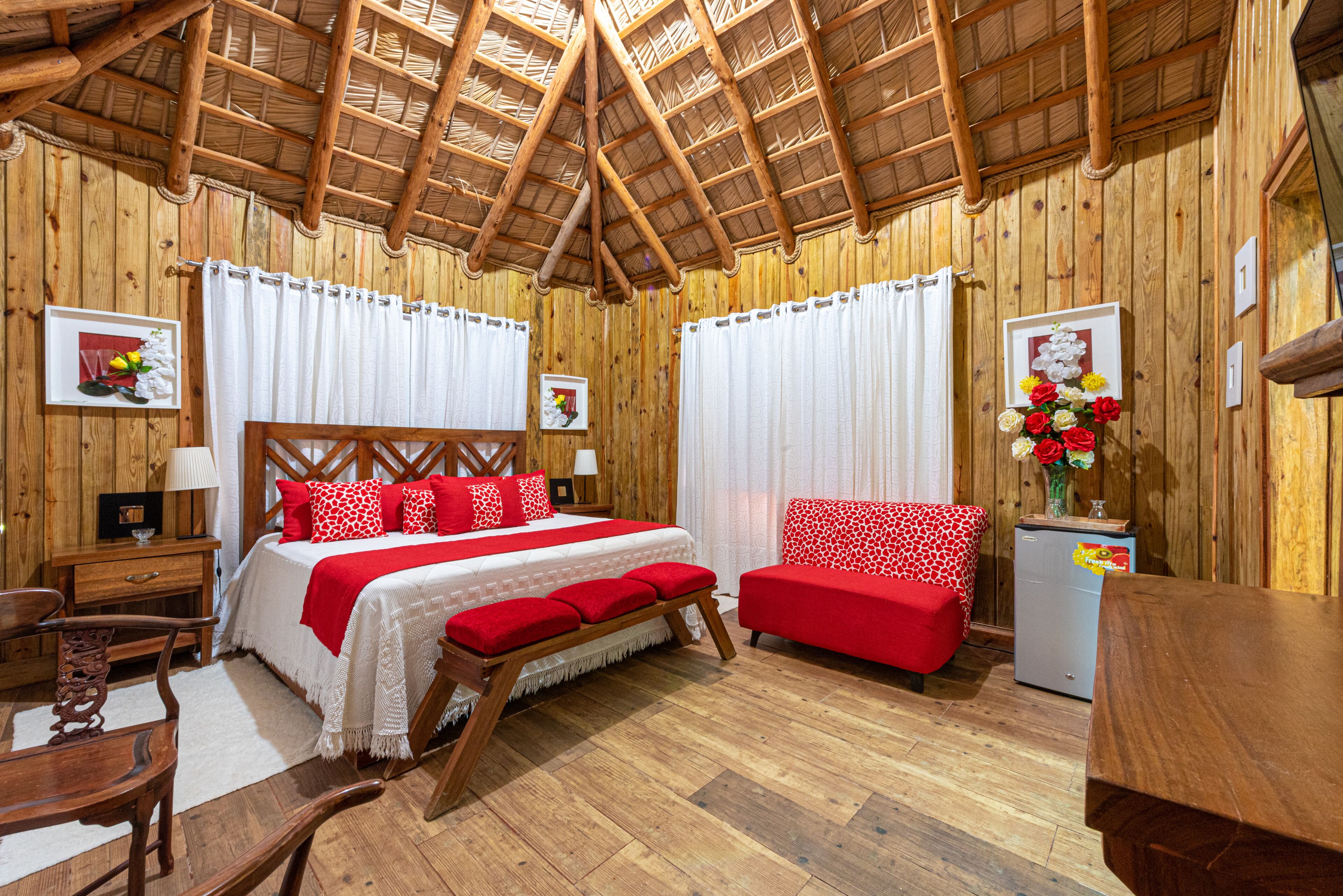 A wooden cabin-style room with a bed, red and white furnishings, a small table, and a refrigerator.