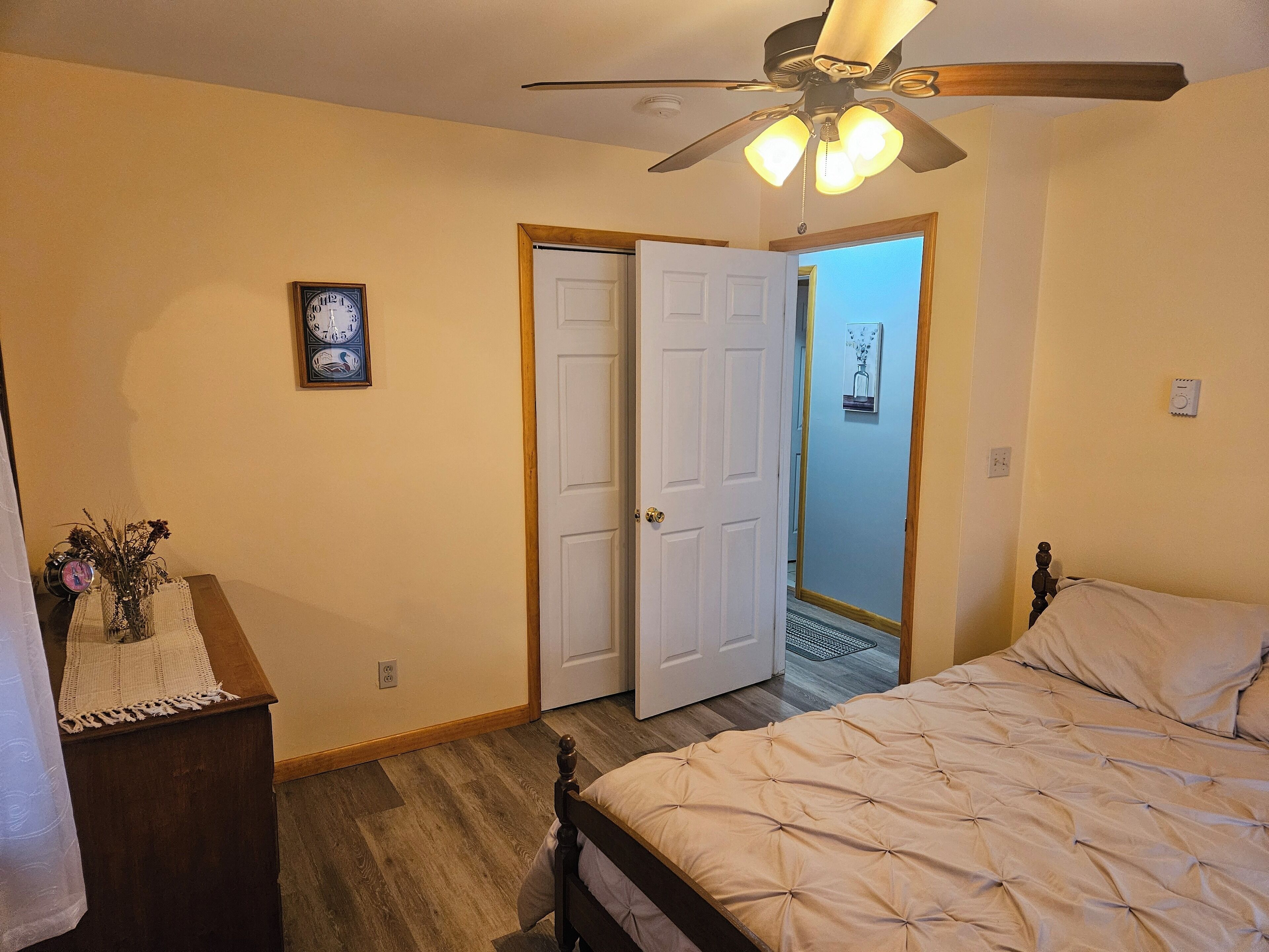 Un dormitorio con una cama, ventilador de techo, una puerta y una pequeña mesa con un reloj y flores.