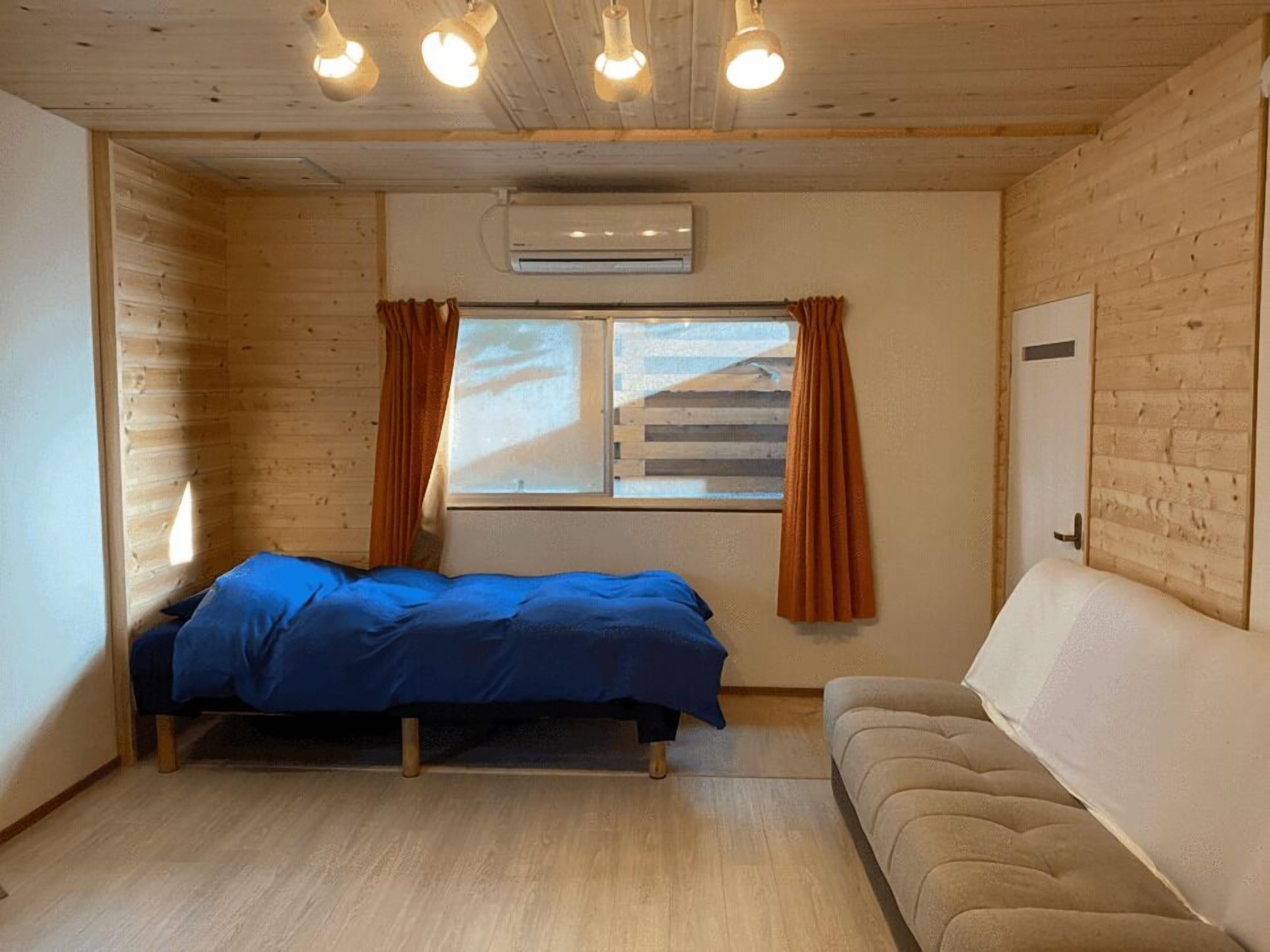 Une pièce comprenant un lit, un canapé, une fenêtre avec des rideaux, un appareil de climatisation et des murs en bois.
