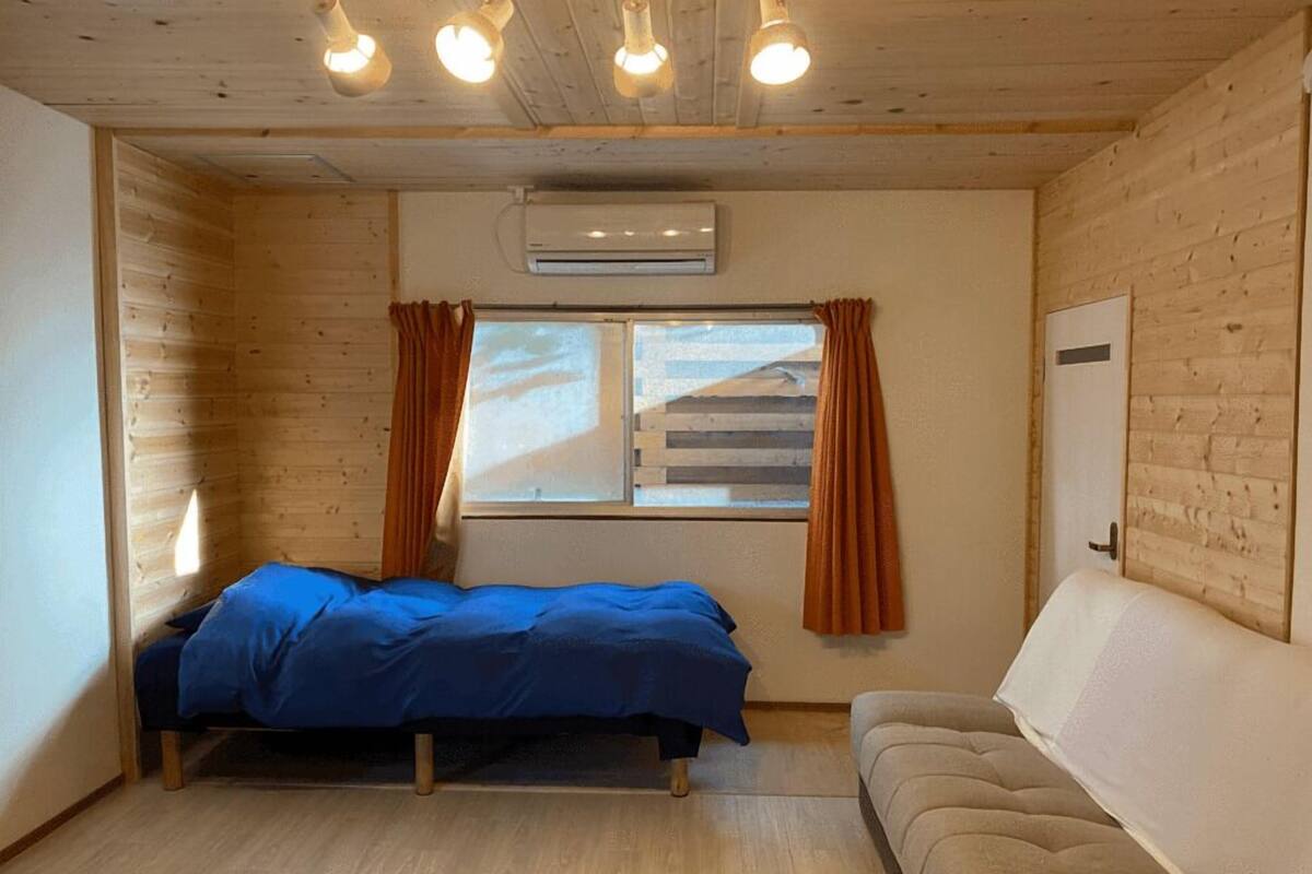 Une pièce comprenant un lit, un canapé, une fenêtre avec des rideaux, un appareil de climatisation et des murs en bois.