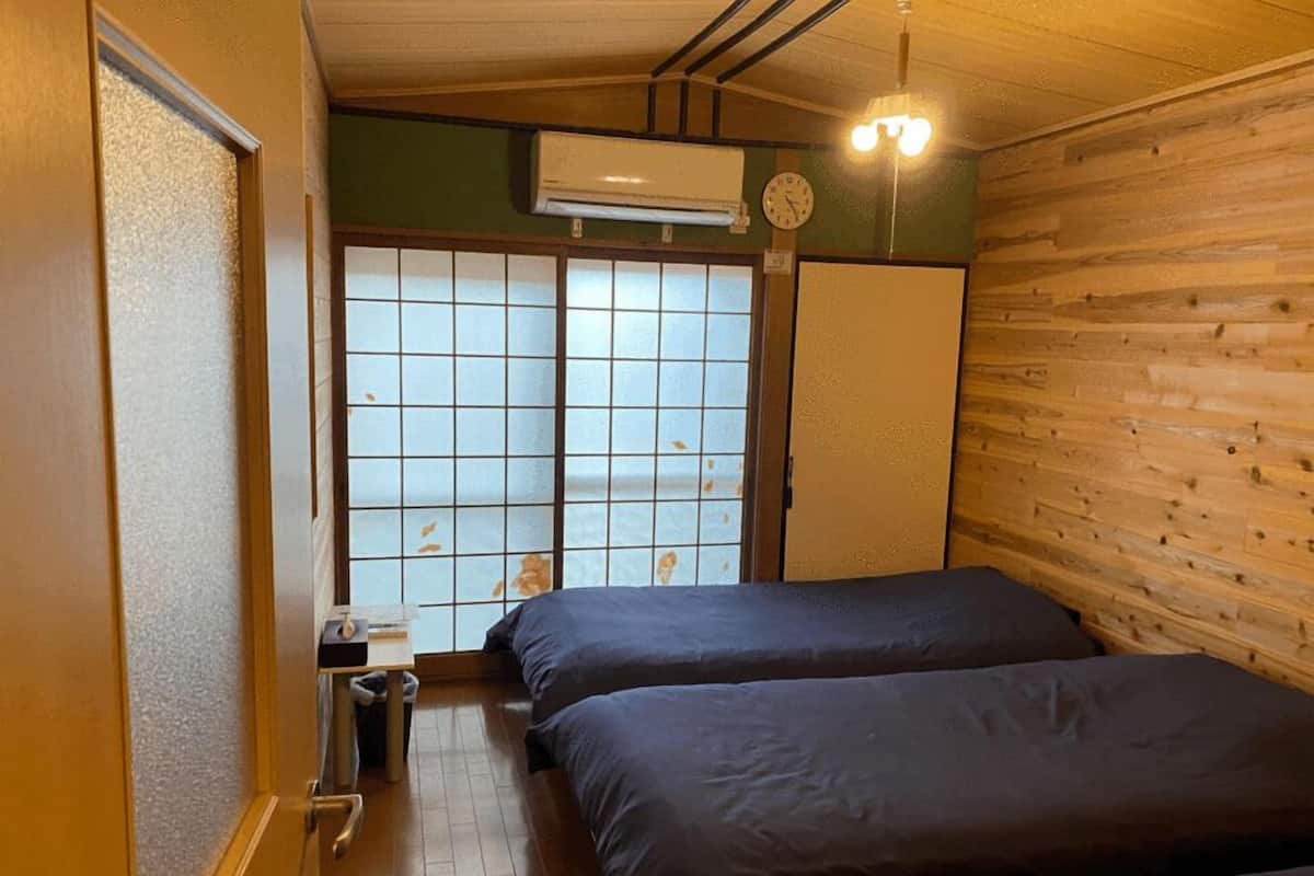 A room with two beds, a wooden wall, a window with a grid pattern, and a wall-mounted air conditioner.