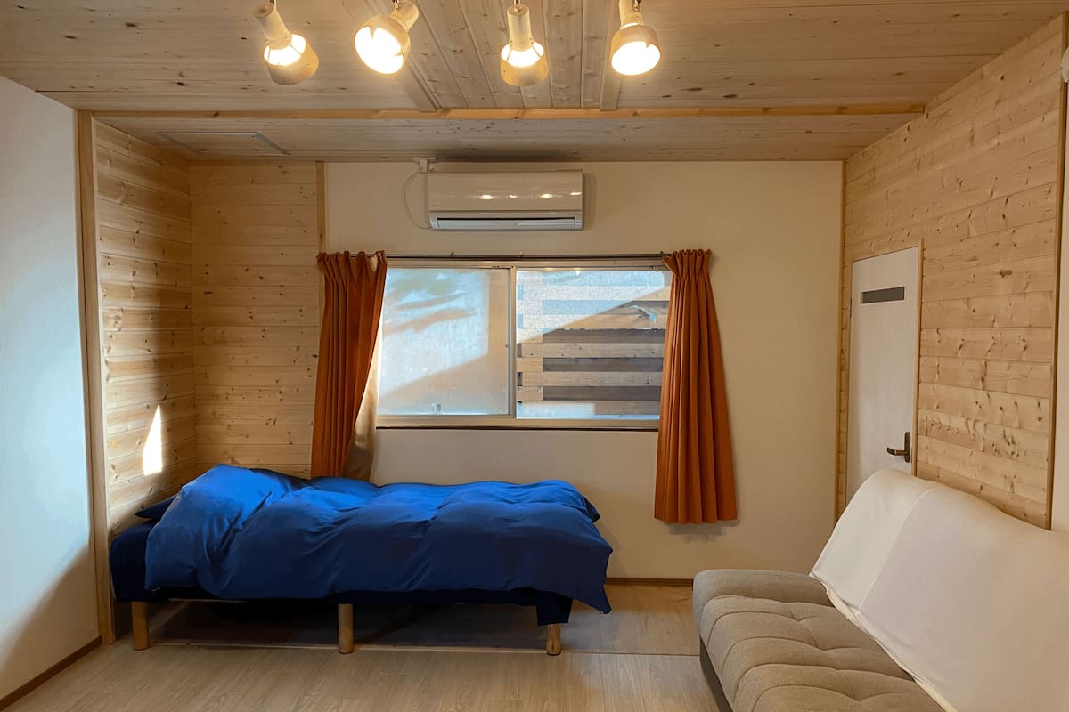 A room with a bed, a sofa, wooden walls, a window with curtains, and an air conditioning unit.
