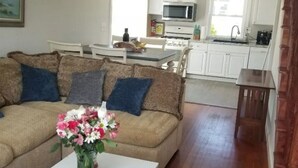 Ein Wohnzimmer mit einem beigefarbenen Sofa, einem weißen Couchtisch und einem Blumenstrauß. Im Hintergrund steht ein Esstisch mit Stühlen und eine Küchenzeile.