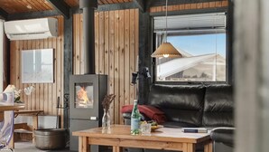 A cozy living room with a wood-paneled wall, a black leather sofa, a wooden coffee table with a bottle and glasses, a fireplace, and a window with a view of a building.