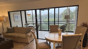 Een woonkamer met een beige bank, een ronde tafel en stoelen. Er is een groot raam met uitzicht op groen en een balkon.