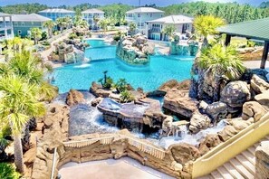 Un complexe hôtelier doté d’une grande piscine extérieure agrémentée de formations rocheuses et de cascades, entouré de palmiers et de bâtiments modernes.