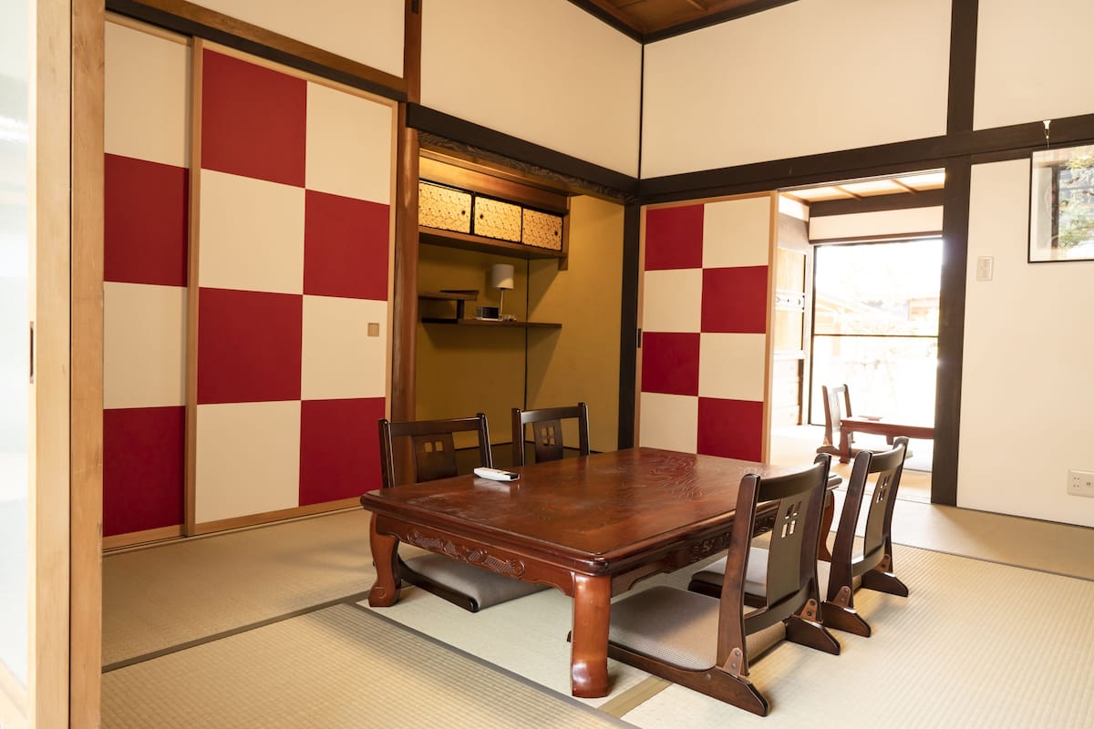 Una habitación tradicional japonesa con suelo de tatami, una mesa de comedor de madera con sillas y puertas correderas shoji con diseños rojo y blanco.