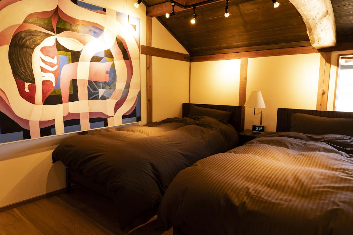 Un dormitorio con un cuadro abstracto grande en la pared, una cama con ropa de cama oscura, una mesita de noche con una lámpara y un techo de madera.