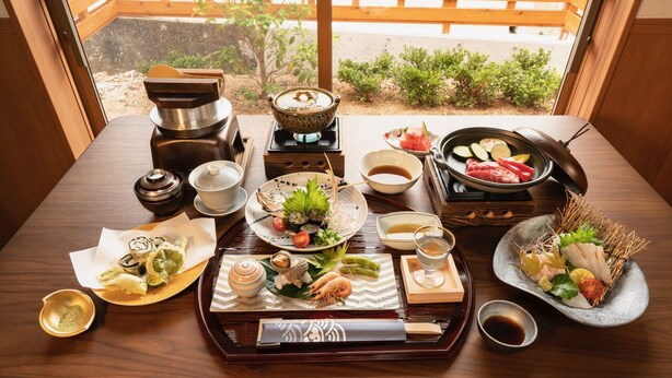 Un montaje tradicional de comida japonesa con varios platos y utensilios sobre una mesa de madera, visto a través de una ventana con una valla de madera afuera.