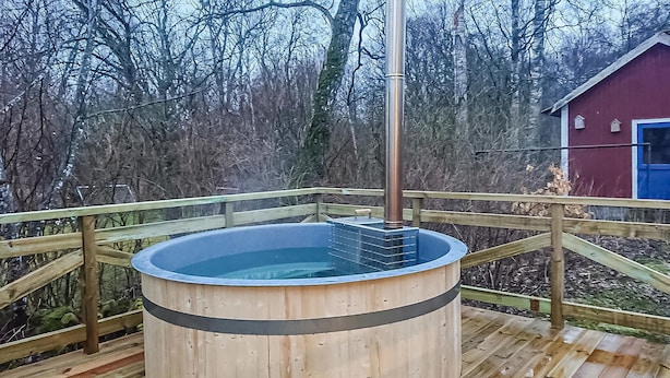 Un jacuzzi de madera en una terraza con un poste de metal, rodeado de árboles y un edificio rojo con detalles azules.