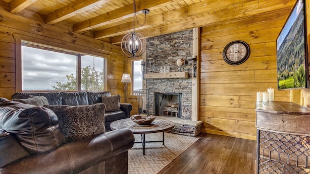 Interior acogedor de una cabaña de madera con un sofá de cuero, una chimenea de piedra y un ventanal con vistas a un área boscosa.
