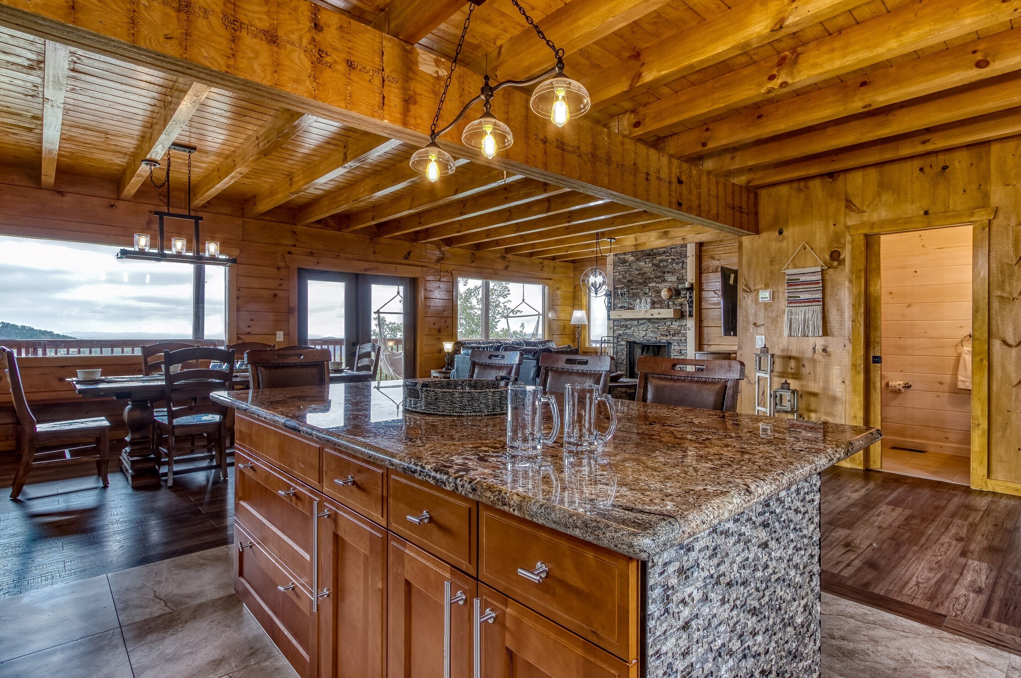 A spacious kitchen with granite countertops, wooden cabinets, and a large island. There are hanging light fixtures and a stone fireplace.